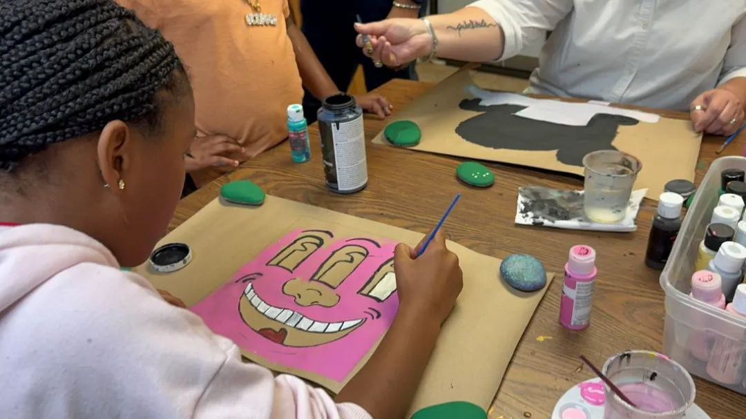 Close-up view of a person painting a large, pink cartoon face with a wide smile onto paper, surrounded by other people painting rocks and using acrylic paints.