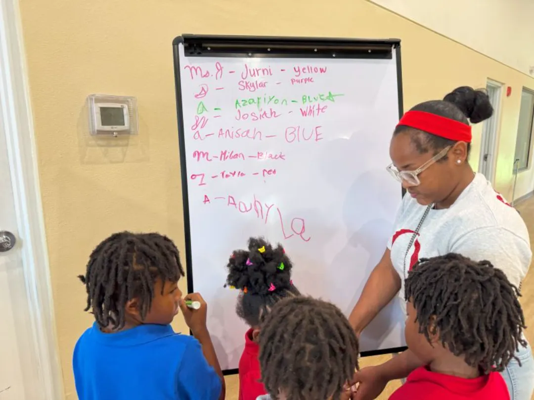 An adult teacher guides three young children, who have dreadlocks, as they write names and corresponding colors on a small whiteboard during a classroom activity