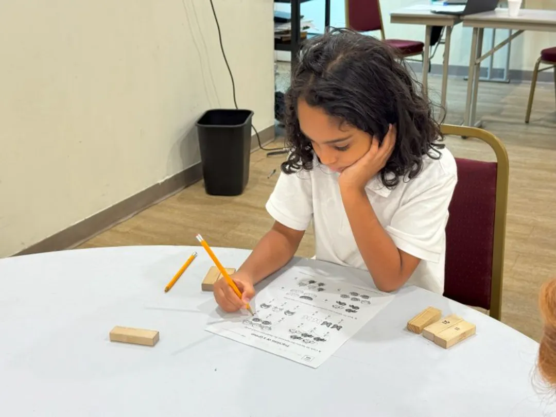 A young student with long dark hair sits at a table, resting her head on her hand while concentrating on a math or counting worksheet with a pencil.