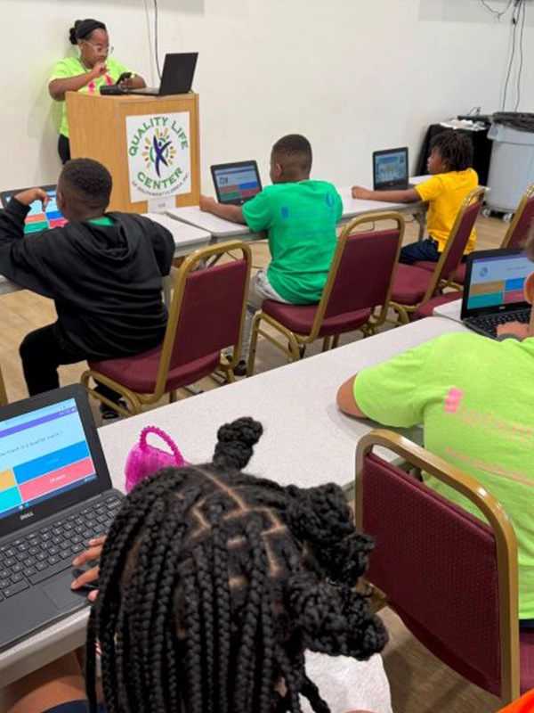 Students participate in an educational activity using laptops, while an adult facilitator stands at a podium displaying the "Quality Life Center" logo in the background.